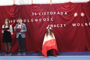 Na czerwonym tle na którym zawieszono biały napis 11 listopada niepodległość znaczy wolność stoi dwójka uczniów prowadzących apel a obok nich siedzi uczennica symbolizująca Polskę - zakrytapolską flagą z biało czerwonym wiankiem na głowie.