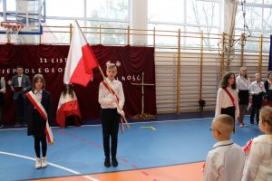 Poczet flagowy podczas odśpiewania hymnu na sali gimnastycznej. Troje uczniów w strojach galowych stoi w odstępach od siebie. Każduy ma przepasaną biało czerwoną wstęgę, a uczeń stojący po środku trzyma flagę. W tle widać drabinki sali, na których zawieszono czerwony materiał a na nim biały napis 11 listopada niepodległość czyli wolność.