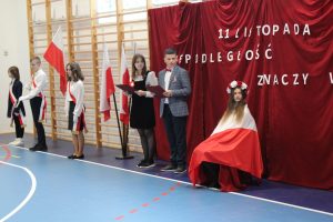 W jednym rzędzie stoją uczniowie z pocztu flagowego szkoły para prowadzących apel oraz na krzesełu siedziuczennica symbolizująca Polskę - zakryta jest białą flaga a na głowie ma wianek z bialych i czerwonych wiatów. W le widać fragment dekoracji - na czerwonym tle napis 11 listopada niepodległość znaczy.