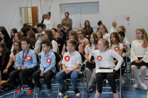 Uczniowie klasy czwartej podczas apelu siedzą na krzesełkach. Dzieci mają przypięte kotyliony i ubrane są na galowo.