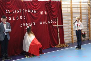 Uczeń stoi z mikrofonem w jednej dłoni i czerwoną różą w drugiej. Przemawia w kierunku uczennicy siedzącej na krzesełku i okrytej biało-czerwoną flagą. Między nimi widać oparty o czerwony materiał krzyż wykonany z dwóch kawałków drzewa. W tle czerwony materiał z napisem 11 listopada wolność znaczy niepodległość.