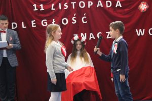 Dwoej uczniów klasy drugiej stoi na przeciwko siebie. Chlopiec trzyma w wyciągniętej dłoni mikrofon. Pomiędzy nimi w lekkim oddaleniu siedzi na krzesełku uczennica okryta biało czerwoną flagą oraz z wiankiem na głowie.