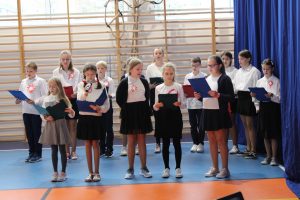 Uczniowie klasy piątej podczas śpiewania piosenek na apelu. Ubrani są w biale koszule a rękach trzymają podkładki z tekstem piosenek.