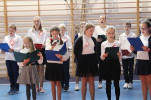 Uczniowie klasy piątej podczas śpiewania piosenek na apelu. Ubrani są w biale koszule a rękach trzymają podkładki z tekstem piosenek.