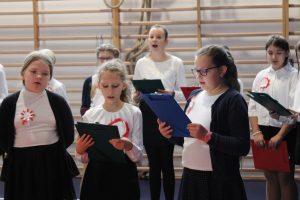 Uczniowie klasy piątej podczas śpiewania piosenek na apelu. Ubrani są w biale koszule a rękach trzymają podkładki z tekstem piosenek.