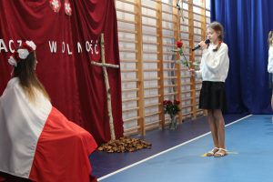 Uczennica stoi na przeciwko siedzącej na krześle koleżanki okrytej biało czerwoną flagą. Dziewczynka przemawia do mikrofonu i trzyma w dłoni czerwoną różę. W tle widać krzyż wykonany z dwóch kawałków drzewa.