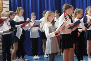 Uczniowie klasy ósmej podczas wykonywania piosenek na apelu z okazji Dnia Niepodległości. Uczniowie stoja obok siebie w rzędach a w dłoniach trzymają podkładki z tekstem piosenek.