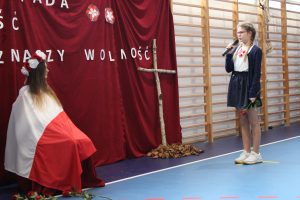 Uczennica z różą w jednej ręce i mikrofonem w drugiej stoi na przeciwko koleżanki siedzącej na krzesełku i okrytej flagą Polski. Między dziewczynkami stoi oparty o czerwony materiał krzyż wykonany z kawałku drzewa. Pod krzyżem leżą uschnięte liście.