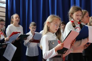 Uczniowie klasy piątej podczas śpiewania piosenek na apelu. Ubrani są w biale koszule a rękach trzymają podkładki z tekstem piosenek.