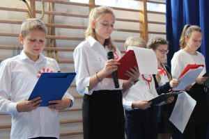 Uczniowie klasy piątej podczas śpiewania piosenek na apelu. Ubrani są w biale koszule a rękach trzymają podkładki z tekstem piosenek.