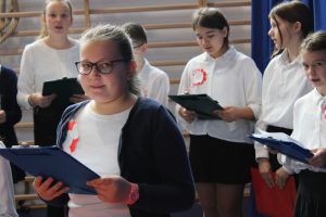 Uczniowie klasy piątej podczas śpiewania piosenek na apelu. Ubrani są w biale koszule a rękach trzymają podkładki z tekstem piosenek. Uczennica stojąca na pierwszym planie lekko uśmiecha się do zdjęcia.