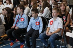 Uczniowie siedzą na ławeczkach podczas apelu z okazji Dnia Niepodległości. Do ubrań maja przyczepione kotyliony.