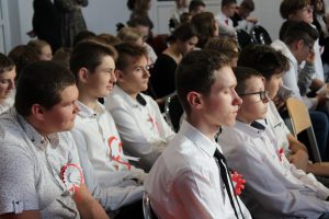 Uczniowie klasy ósmej patrza przed siebie siedząc na krzesełkach podczas apelu z okazji Dnia Niepodległości.