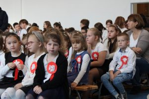 Uczniowie siedzą na ławeczkach podczas apelu z okazji Dnia Niepodległości. Do ubrań maja przyczepione kotyliony.