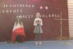 Na czerwonym tle z napisem 11 listopada niepodległość znaczy wolność, siedzi uczennica okryta biało-czerwoną flagą. Przed nią z prawej strony stoi uczennica klasy piątej z mikrofonem w dłoniach. Na dziewczynki padają promienie sloneczne.