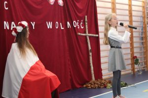 Uczennica klasy czwartej stoi bokiem do zdjęcia i spiewa do mikrofonu. Za nią siedzi dziewczynka okryta Polską flagą. W tle widać oparty o czerwony materiał krzyż z kwałków drzewa.