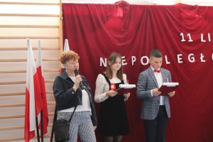 Na czerownym tle obok flag Państwowych stoją Pani Wiesława urbańska ora dwoje uczniów prowadzących apel z okazji Dnia niepodległosci. Pani Wiesława przemawia przez mikrofon a uczniowie trzymają w dłoniach czerwone podkładki.