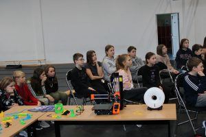 Uczniowie siedzą na krzesełkach na sali gimnastycznej. Przed nimi na ławce stoi drukarka 3D oraz kolorowe przedmioty przez nią wykonane.
