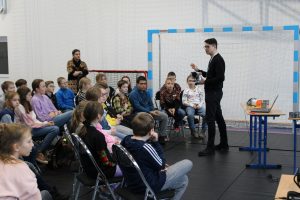 Pan prowadzący zajęcia z mobilnego laboratorium stoi przed uczniami siedzącymi na krzesełkach. Mężczyzna trzyma w dłoniach gogle.