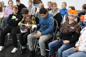 Uczniowie siedzą na krzesełkach na sali gimnastycznej. Dwóch chłopców siedzących w pierwszym rzędzie ma założone gogle Vr a dwójka siedząca obok jest w trakcie ich zakładania.