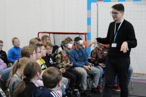 Pan prowadzący warsztaty z mobilnego laboratorium stoi przed uczniami ze zgiętymi w łokciu rękami i opuszczonymi dłońmi. Dzieci patrzą na niego. Troje uczniów w pierwszym rzędzie ma założone gogle VR.