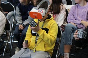 Uczeń siedzi na krzesełku. W tle widać siedzące inne dzieci. Chłopiec ma założone gogle Vr a w dłoniach trzyma kontroler oraz czarnobiałą kostkę.