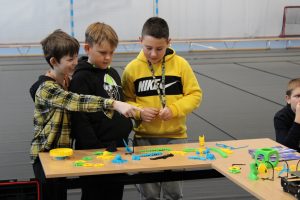 Trzech uczniów stoi przy stoliku, na którym rozłożone są rzeczy wydrukowane z drukarki #D. Jeden z chłopców smiejąc się pokazuje coś wyciągnięta dłonią swoim kolegą.