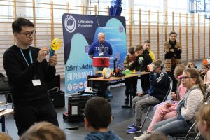 Pan prowadzący warsztaty z mobilnego labolatorium trzyma w dłoniach wiatrak wydrukowany na drukarce 3D. Naprzeciwko siedzą uczniowie a z boku stoi stolik z drukarką i wydrukowanymi na niej przedmiotami.