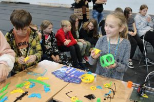 Uczennica stoi przy stoliku z wydrukowanymi z drukarki 3D przedmiotami. Dziewczynka trzyma w dłoniach dwa z nich i się uśmiecha. Obok stoi jej kolega również przyglądający się przedmiotom. W tle widać siedzących na krzesełkach uczniów.