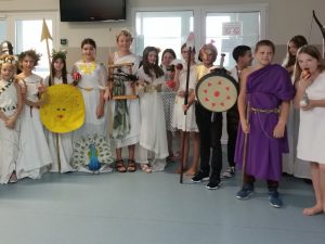 Uczniowie klasy piatej stoją w półkolu obok siebie. Dzieci przebrane są za greckich bogów i boiginie. Dziewczynki ubrane są w białe toki z wiankami na glowie. Jeden chłopiec przebrany jest za greckiego wojownika, a jego kolega ma założoną fioletową togę.