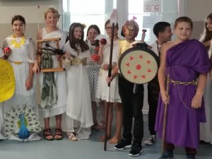 Uczniowie klasy piatej stoją w półkolu obok siebie. Dzieci przebrane są za greckich bogów i boiginie. Dziewczynki ubrane są w białe toki z wiankami na glowie. Jeden chłopiec przebrany jest za greckiego wojownika, a jego kolega ma założoną fioletową togę.