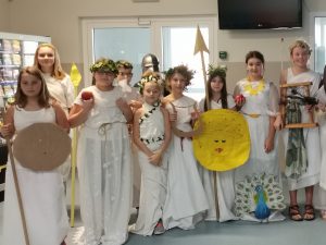 Uczennice klasy piątej pozują do zdjęcia grupowego w strojach przedstawiających greckie boginie. Dziewczynki trzymaja atrybuty swoich boigini takie jak tarcze i jabłka. Na głowach mają wianki.