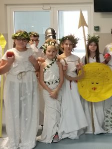 Uczennice klasy piątej pozują do zdjęcia grupowego w strojach przedstawiających greckie boginie. Dziewczynki trzymaja atrybuty swoich boigini takie jak tarcze i jabłka. Na głowach mają wianki.