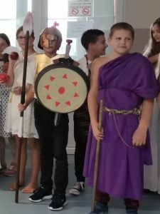 Dwóch uczniów klasy piątej stoi obok siebie w przebraniu greckich wojowników. Jeden z nich ma na głowie hełmn a wręku trzyma włócznię i tarczę. Drugi zaś ma założoną fioletową togę.