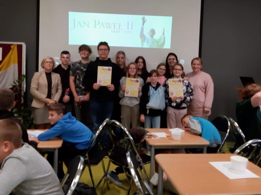 Uczniowie wraz z Panią Dyrektor oraz dwie nauczycielkami pozują do zdjęcia grupowego. Troje uczniów trzyma przed sobą dyplomy. Wszyscy stoją na tle ściany, na której wyświetla się napis JAN PAWEŁ II oraz podobizna papieża. Przed fotografowaną grupą przy stolikach siedzą pozostali uczniowie.