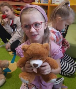 Dziewczynka z oddziału przedszkolnego trzyma wrekach swojego pluszowego misia. W tle widać inne dzieci z maskotkami.