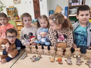 Dzieci z oddziału przedszkolnego stoją wokół stolika na którym ustawiono w rządku modele misi. Przed modelami leżą złożone przez uczniów inne modele misi. Część dzieci trzyma swoje pluszowe misie.