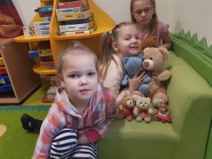 Trzy dziewczynki z oddziału przedszkolnego siedzą przy zielonej kanapie, na której ułożone są pluszowe misie. Jedna z dziewczynek się do nich przytula.