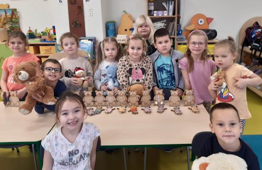 Dzieci z oddziału przedszkolnego stoją wokół stolika na którym ustawiono w rządku modele misi. Przed modelami leżą złożone przez uczniów inne modele misi. Część dzieci trzyma swoje pluszowe misie.