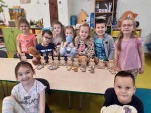 Dzieci z oddziału przedszkolnego stoją wokół stolika na którym ustawiono w rządku modele misi. Przed modelami leżą złożone przez uczniów inne modele misi. Część dzieci trzyma swoje pluszowe misie.