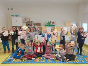 Uczniowie klasy pierwszej pozują do grupowego zdjęcia. Część dzieci stoi część siedzi na dywanie. Wszystkie dzieci podnoszą wycięte z papieru i pokolorowane misie.