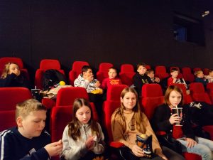 Uczniowie klasy piatej siedzą w czerwonych fotelach na sali kinowej. Dzieci maja popcorn i napoje.