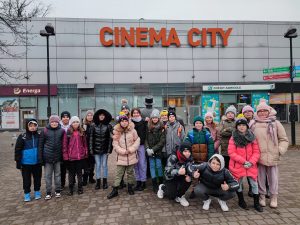 Uczniowie klasy piatej pozują do grupowego zdjęcia przed budynkiem cinema city. Nad dziećmi widoczny jest napis budynku.
