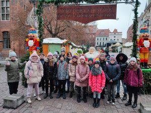 Uczniowie klasy piątej pozują do wspólnego zdjęcia pod napisem Toruński jarmark bożonarodzeniowy. Za nimi widać postać sztucznego mikołaja o po bokach dwóch żołnierzyków z dziadka do orzechów.
