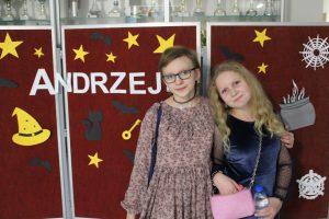 Dwie uczennice klasy czwartej pozują do wspólnego zdjęcia na tle czerwonej tablicy z napisem "ANDRZEJKI". Jedna z dziewczynek obejmuje ramieniem drugą. Obie ubrane w sukienki.
