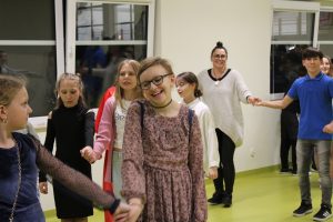 Uczniowie tańczą w parach na zabawie andrzejkowej. Dziewczynka stojąca na pierwszym planie szeroko się uśmiecha. W tle widać ucznia w niebieskiej koszuli tańczącego ze swoją wychowawczynią.
