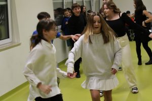 Uczniowie tańczą w parach podczas zabawy adrzejkowej. W pierwszej parze dwie uczennice ubrane na biało trzymają się za ręce.