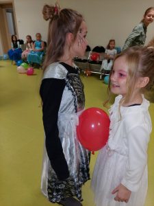 Dwie uczennice stoją naprzeciwko siebie w przebraniach. Pomiędzy nimi utrzymuje się czerwony balonik.