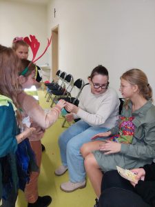 Dwie uczennice z klasy szóstej siedzą na krzesełkach pod ścianą. Jedna z nich przekazuje cukierka dziewczynce stojącej przed nią.