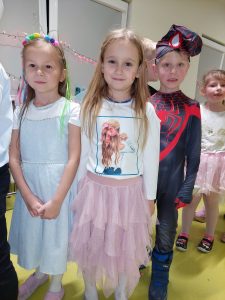 Dwie dziewczynki stoją w jasnych sukienkach obok siebie. Za nimi stoi chłopiec przebrany za spidermena.
